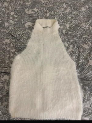 Vit stickad ärmlös polotröja - Säljer en fluffig, vit stickad ärmlös polotröja i mjukt material. Tröjan har hög krage och rak passform, perfekt för lager på lager och trendiga outfits. Passar dig som gillar minimalistisk och stilren look.