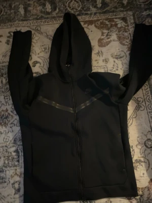 Nike teech fleece hoodie - Säljer en svart zip hoodie från Nike med huva och hel dragkedja. Tröjan har diskreta svarta detaljer över bröstet och två fickor framtill. Perfekt för träning eller chill. Klassisk Nike-logga på bröstet och mjukt material.