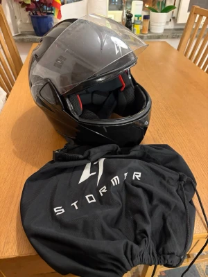 MC hjälm - Säljer en svart mc-hjälm från Stormer med genomskinligt visir och röd knapp på insidan som öppnar främre hjälmen. Hjälmen är aerodynamiskt formad och kommer med en svart tygpåse med Stormer-logga. Finns några tecken på användning som skrap märken och revor lite här och där men funkar fortfarande som ny! Har bara använts en sommar säsong:) storlek S. 