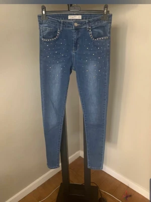 Blå jeans med nitar från Hello Miss - Snygga blå jeans från Hello Miss med smal passform och dekorativa silverfärgade nitar framtill. Jeansen har klassisk femficksdesign och är tillverkade i stretchigt denimtyg för extra komfort. Perfekta för dig som vill ha en cool och trendig look.
