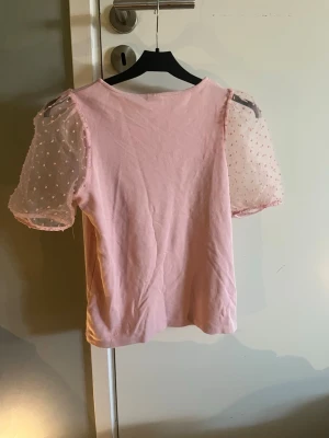 Rosa topp med puffärmar från Lindex - Supersöt rosa topp från Lindex med transparenta puffärmar i mesh och små prickar. Kroppen är i mjuk bomull och ärmarna ger en extra gullig touch. Perfekt för dig som vill ha något unikt och trendigt i garderoben!