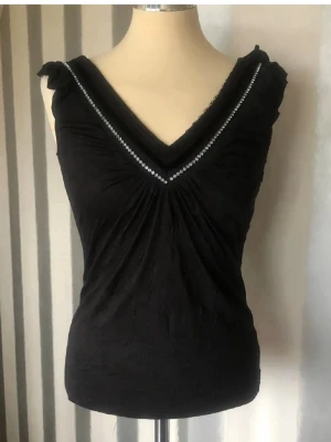 Svart ärmlös topp med pärldetaljer - Snygg svart ärmlös topp med djup v-ringning och pärldetaljer längs urringningen. Toppen har lätt rynkade axlar och en smickrande passform. Perfekt för dig som vill ha en stilren men ändå glittrig look.
