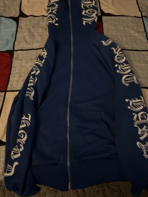 Blå hoodie med vita tryck - Blå hoodie med dragkedja och stora vita tryck längs båda ärmarna och huvan. Hoodien har en klassisk passform, ribbade muddar och en stor huva. Materialet ser ut att vara mjukt och bekvämt, perfekt för en avslappnad stil.