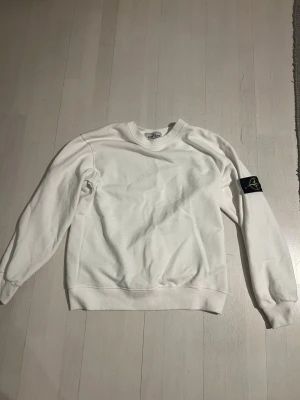 Stone Island sweatshirt  - Vit sweatshirt från Stone Island med klassisk rund hals och ribbade muddar. Tröjan har den ikoniska svarta logotyp-patchen på vänster ärm. Perfekt för en clean och stilren look.