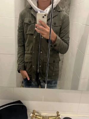 Olivgrön utilityjacka från j lindeberg - Snygg olivgrön fieldjacket från J Lindeberg med fyra stora fickor framtill och knappar. Jackan har dragkedja och knappstängning, klassisk krage och är tillverkad i ett slitstarkt bomullsmaterial. Perfekt för lager-på-lager