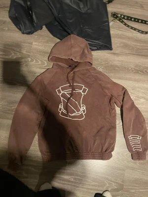 Number (N)ine Shield hoodie - Nettspend hoodie Säljer en brun hoodie från number (n(ine med huva och snörning. Den har ett stort vitt grafiskt tryck på framsidan och ett mindre tryck på ena ärmen. Hoodien har ribbade muddar och är tillverkad i ett mjukt material, perfekt för en avslappnad och cool look.