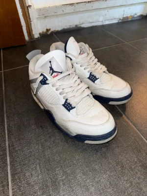 Vita Air Jordan 4 Retro sneakers - Säljer ett par vita Air Jordan 4 Retro sneakers med marinblå och grå detaljer. Skorna har klassisk mesh på sidorna, Jumpman-logga på plösen och robust gummisula. Perfekta för dig som gillar streetwear och ikoniska modeller.