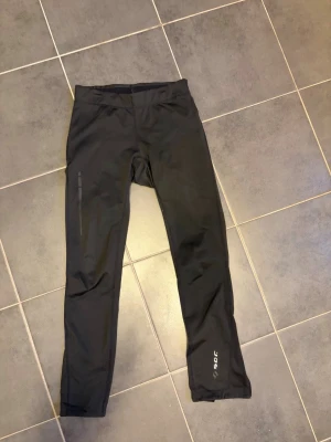 Svarta träningsbyxor från SOC - Svarta träningsbyxor från SOC med smal passform och dragkedjor vid bensluten. Byxorna har en diskret logga på ena benet och är tillverkade i ett stretchigt funktionsmaterial som passar perfekt för löpning eller gym. Har en ficka på baksidan/ sidan. Uppskattning storlek xs-s