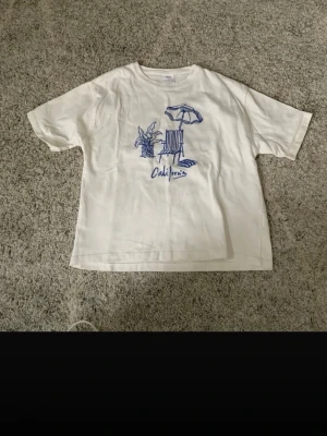 Vit t-shirt med blå California-tryck - Vit t-shirt med blått tryck som föreställer en strandstol, parasoll och palm samt texten 'California' på framsidan. T-shirten har en avslappnad passform och korta ärmar, perfekt för en chill look.