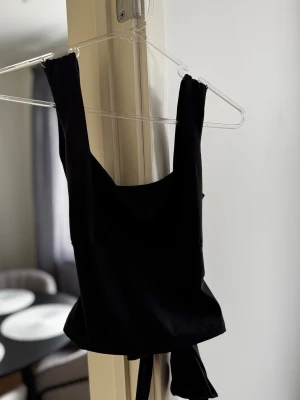 Svart crop top från DesignBySi - Trendig svart crop top från DesignBySi med breda axelband och knytband i ryggen. Toppen ett stretchigt, mjukt material som sitter snyggt på kroppen. Perfekt för att skapa en cool och stilren look. Prislapp kvar