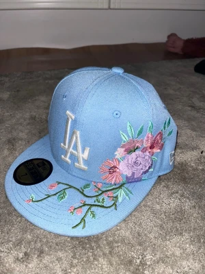 Blå LA-keps med blommigt broderi - Säljer en ljusblå New Era 59FIFTY keps med LA-logga framtill och snyggt broderade blommor i rosa, lila och grönt på sidan och skärmen. Kepsen har rak skärm och klassisk passform. Perfekt för dig som vill sticka ut med en unik accessoar.