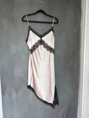 Spets klänningen  - Snygg klänning i champagnefärgad satin med svarta spetsdetaljer längs urringning, midja och nederkant. Asymmetrisk fåll och tunna axelband ger en elegant och modern look. Perfekt för dig som vill känna dig extra lyxig på kvällen. Helt helt ny!