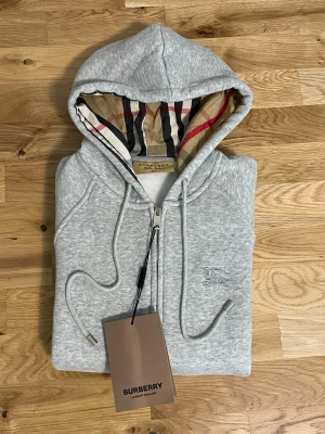 Zip hoodie - Snygg grå zip hoodie med klassiskt rutig foder i huvan och diskret broderad logga på bröstet. Hoodien har dragsko i huvan, ribbade muddar och fickor framtill. Tillverkad i mjuk bomullsblandning för extra komfort.