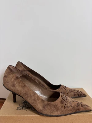 Bruna mockapumps med broderi - Snygga bruna pumps i mocka med spetsig tå och låg klack. Skorna har ett unikt blommigt broderi på tån som ger en cool vintagekänsla. Perfekta för dig som vill ha något extra till outfiten. Använda fåtal gånger!💞