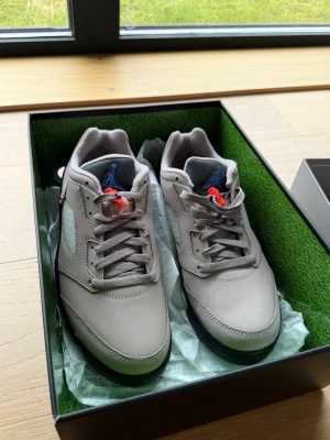 Jordan x PSG grå Air jordan 5 Low - Snygga Jordan x Paris Saint-Germain sneakers i grå mocka med svarta och röda detaljer. Lågt skaft, meshpaneler på sidorna och blå Jumpman-logga på plösen. Sulan är svart och grå, och skorna har en unik PSG-detalj vid hälen. Perfekta för dig som gillar streetwear.