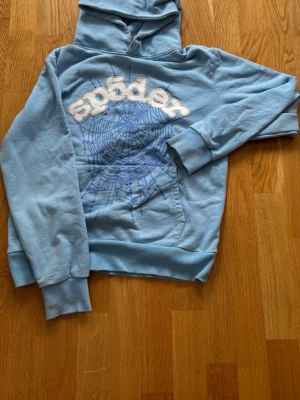 Ljusblå hoodie från Sp5der - Säljer en ljusblå hoodie från Spyder med stor vit logga och ett coolt spindelnätsmönster på framsidan. Hoodien har huva, ribbade muddar och en klassisk magficka. Perfekt för dig som gillar streetwear och vill sticka ut lite extra.