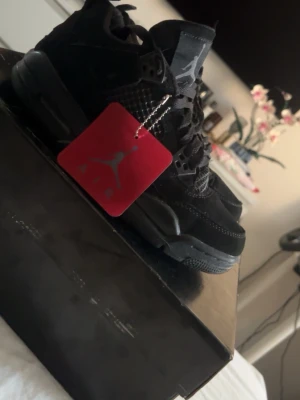 Svarta Air Jordan 4 Retro sneakers - Snygga helsvarta Air Jordan 4 Retro sneakers med meshdetaljer och klassisk Jumpman-tag. Skorna har en robust sula och ikoniska plastdetaljer vid snörningen. Perfekta för dig som gillar streetwear och vill ha en stilren look.