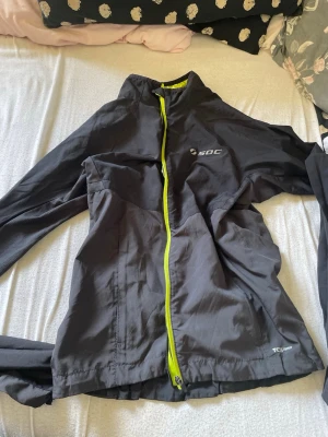 Svart vindjacka från SOC - Snygg svart vindjacka från SOC med neongul dragkedja och detaljer. Jackan har hög krage, justerbar nederkant och är tillverkad i ett lätt, vindtätt material. Perfekt för utomhusaktiviteter och sportiga dagar.