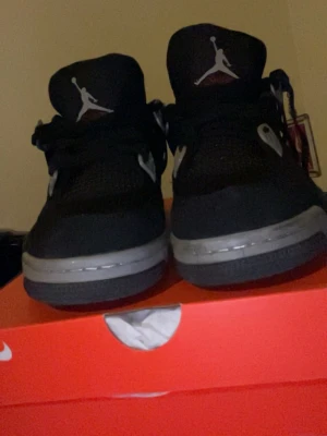 Svarta Air Jordan 4 Retro sneakers - Säljer ett par svarta Air Jordan 4 Retro sneakers med vita och grå detaljer. Skorna har meshpaneler, synlig Air-enhet i sulan och Jumpman-logga på plösen. Klassisk siluett med robust känsla och cool design. Perfekta för dig som gillar streetwear och sneakerskultur.