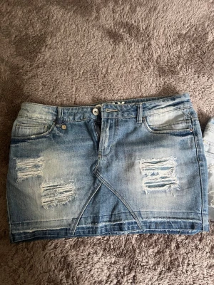 Blå jeanskjol från ONLY - Säljer en snygg blå jeanskjol från ONLY med slitna detaljer och fransar framtill. Kjolen har klassisk femficksdesign, bälteshällor och knappgylf. Perfekt för dig som gillar en avslappnad och trendig stil.