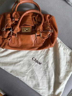 Chloé Paddington väska - Säljer en snygg brun handväska från Chloé i mjukt läder med guldfärgade metalldetaljer och dekorativa nitar. Väskan har dubbla handtag, låsdetalj med nyckel och kommer med original dustbag. Perfekt för dig som gillar lyxiga accessoarer.