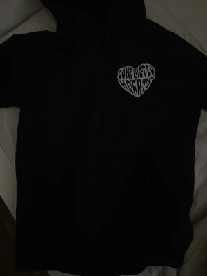 Svart hoodie Conflicted Hearts - Säljer en svart hoodie från Conflicted Hearts med vit text i form av ett hjärta på bröstet. Klassisk modell med huva och dragsnören. Perfekt för dig som gillar streetwear och vill ha en enkel men snygg hoodie.
