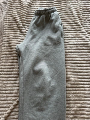 Gråa mjukisbyxor från Nike - Säljer ett par gråa mjukisbyxor från Nike med diskret logga på benet. Byxorna har resår i midjan och raka ben. Tillverkade i mjukt och bekvämt material, perfekta för chill dagar eller träning.