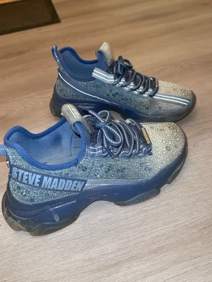Blå glittriga sneakers Steve Madden - Snygga blå sneakers från Steve Madden med glittriga stenar över hela ovandelen. Skorna har tjock sula, matchande blå snören och detaljer i silver. Märkeslogga på sidan och plös. Perfekta för dig som vill sticka ut med din stil.