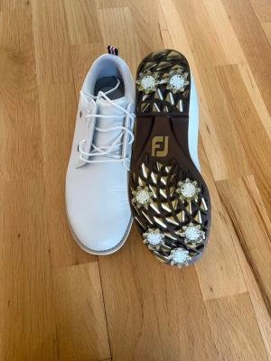 Vita golfskor från FootJoy Premiere - Snygga vita golfskor från FootJoy med klassisk design och snörning. Skorna har en ovandel i slittåligt syntetmaterial och greppsäker sula med utbytbara softspikes. Perfekta för golfbanan och ger bra stabilitet och komfort under rundan.