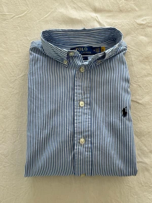 Blårandig skjorta från Polo Ralph Lauren - Klassisk blårandig skjorta från Polo Ralph Lauren med button-down krage och broderad logga på bröstet. Skjortan har vita knappar och lång ärmar. Tillverkad i mjuk bomull för en bekväm känsla och snygg passform.