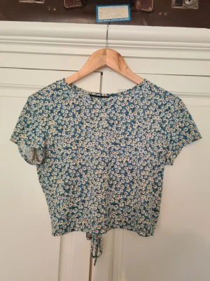 Blommig croppad topp med knyt - Söt blommig topp i blått och vitt med korta ärmar och croppad passform. Toppen har ett knytband i midjan och en öppen detalj i ryggen. För mer exakt färg se den sista bilden. Perfekt för sommarens alla tillfällen. Storlek M/L. Hör gärna av dig vid frågor😁