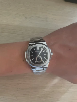 Silverfärgad klocka från MAIMASHI  - Stilren klocka från MAIMASHI med silverfärgad länk i metall och svart urtavla. Klockan har romerska siffror, en mindre sekundvisare och en klassisk rund form. Perfekt accessoar för dig som gillar en clean och modern look.