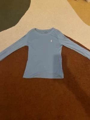 Ljusblå långärmad topp Polo Ralph Lauren - Snygg ljusblå långärmad topp från Polo Ralph Lauren med rund halsringning och den klassiska vita logon broderad på bröstet. Tillverkad i mjukt bomullsmaterial som känns skönt mot huden. Perfekt för en avslappnad och stilren look.                       Skriv om du har några frågor!