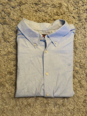 Ljusblå skjorta med broderad detalj - Klassisk ljusblå skjorta från Morris med button-down krage och vita knappar. Skjortan har en liten broderad logga på bröstet och är tillverkad i bomull. Perfekt för en clean och stilren look.