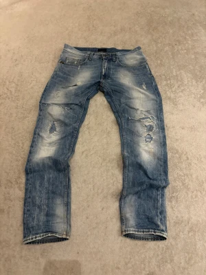 Blåa Tiger of sweden jeans med unika slitningar  - Säljer ett par blå jeans med slitningar och hål på benen. Jeansen har en tvättad look med ljusa partier och detaljerade lagningar. Klassisk femficksmodell och raka ben. Perfekta för en avslappnad och trendig stil. Riktigt unika slitningar!