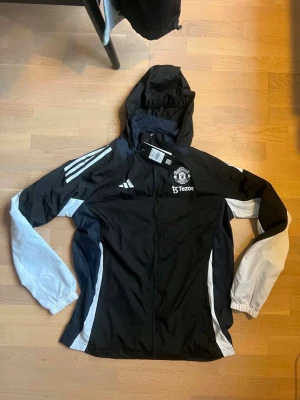 Manchester United vindjacka svart/vit - Snygg svart och vit vindjacka från Adidas med Manchester United-logga och Tezos-tryck på bröstet. Jackan har huva, dragkedja framtill och vita detaljer på ärmar och sidor. Perfekt för dig som vill ha sportig stil och visa ditt lag.