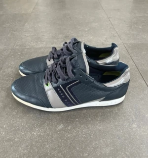 Hugo Boss skor - Snygga mörkblå sneakers från Hugo Boss med grå och vita detaljer. Skorna har snörning, perforerat läder framtill och en vit sula. Märkeslogga på sidan och insidan har gröna detaljer. Använda vid ett tillfälle, som nyttskick