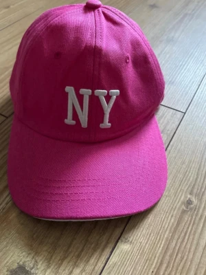 Rosa  keps med böjd skärm - Snygg rosa keps med böjd skärm och broderad vit NY-logga framtill. Klassisk sexpaneldesign och justerbar rem bak. Perfekt för dig som vill sticka ut med en färgstark accessoar. Tillverkad i bomullstyg.