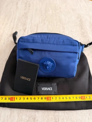  Versace - Snygg blå handväska från Versace i nylon med klassisk Medusa-detalj framtill. Väskan har dragkedja upptill, justerbar axelrem och kommer med originalpåse och etikett. Perfekt för dig som vill ha en stilren och trendig accessoar.