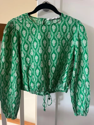 Grön mönstrad blus från Zara - Grön blus från Zara med vitt bladliknande mönster. Blusen har rund halsringning, långa ärmar med lätt puff och dragsko i midjan för en snygg passform. Tillverkad i ett lätt tyg som känns luftigt och skönt.