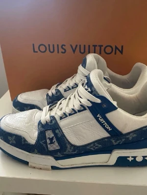 Louis Vuitton blåvita sneakers - Säljer ett par exklusiva Louis Vuitton sneakers i vitt och blått med klassiskt monogrammönster. Skorna har vita läderpaneler, blå denimdetaljer och tydliga LV-loggor på plös och sida. Snygga snörningar och robust sula med stjärndetaljer. 