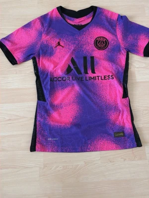 PSG matchtröja rosa/lila Jordan - Säljer en Paris Saint-Germain matchtröja i samarbete med Jordan. Tröjan har ett fett rosa och lila mönster, svart PSG-logga på bröstet och svart Jordan-logga. Tillverkad i lätt och ventilerande material, perfekt för fotboll eller träning.