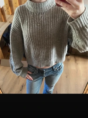 Grå croppad stickad tröja - Säljer en grå, croppad stickad tröja med hög hals och breda ärmar. Tröjan har ett grovt stickat mönster och ribbade muddar vid hals, ärmslut och nederkant. Perfekt för lager på lager och chill stil. Bilderna är från min vinted💕💕