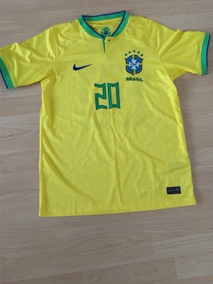 Brasilien matchtröja Nike gul - Säljer en gul Brasilien matchtröja från Nike med gröna detaljer och nummer 20 på bröstet. Tröjan har CBF-emblemet och fem stjärnor ovanför, samt blå Nike-logga. Tillverkad i lätt och ventilerande material, perfekt för fotboll eller träning.