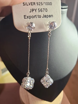 Silverörhängen med kubformade stenar - Säljer ett par eleganta örhängen i silver 925 med långa kedjor och gnistrande kubformade stenar i ändarna. Örhängena har även en rund klar sten vid fästet och ger ett lyxigt intryck med sin moderna design.