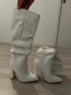 Vita höga boots med klack - Säljer ett par vita höga stövlar med chunky klack och spetsig tå. Stövlarna har en slouchy passform och är tillverkade i ett glansigt syntetmaterial som ger en trendig look. Perfekta för att lyfta vilken outfit som helst.
