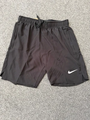 Svarta träningsshorts från Nike - Säljer ett par svarta träningsshorts från Nike med snörning i midjan och elastisk linning med Nike-logga. Shortsen har en vit Nike Swoosh på benet och är tillverkade i ett lätt och snabbtorkande material. Perfekta för gymmet eller löprundan.