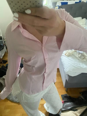 Rosa och vit randig skjorta - Säljer en snygg rosa och vit randig skjorta med klassisk krage och knappar framtill. Skjortan har lång ärm och en figurnära passform som ger en clean och fräsch look. Perfekt att styla till jeans eller kjol för en trendig outfit.