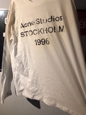 Vit långärmad t-shirt Acne Studios - Vit långärmad t-shirt från Acne Studios med svart tryck på bröstet där det står 'Acne Studios STOCKHOLM 1996'. Klassisk passform och rund halsringning, perfekt för en clean och stilren look.