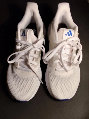 Vita Adidas sneakers med mesh - Fräscha vita sneakers från Adidas med ventilerande mesh-ovandel och klassiska tre ränder på sidan. Skorna har snörning och låg profil, samt blå Adidas-logga på plösen. Perfekta för en sportig och stilren look.